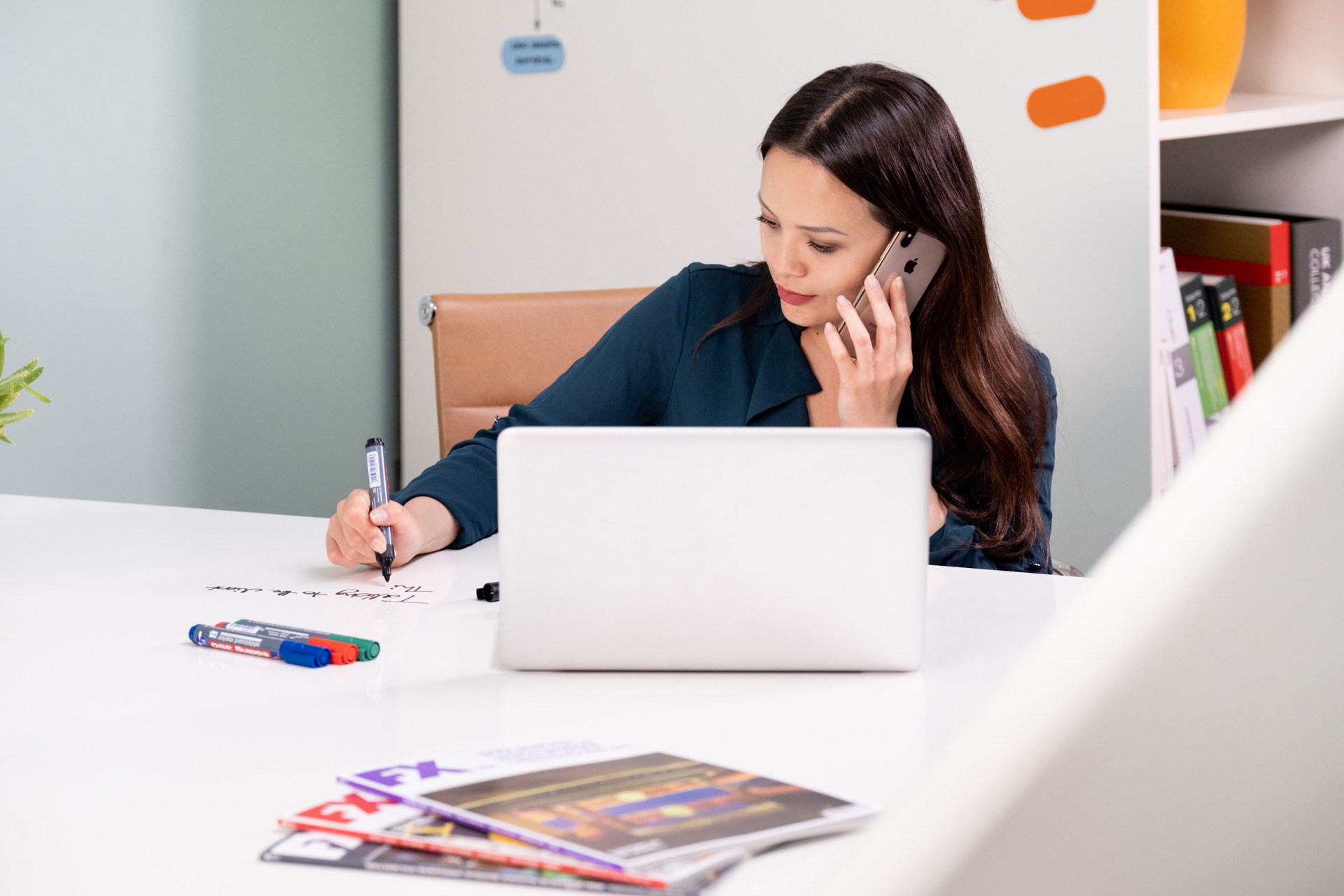 Female office worker on her mobile phone working at a whiteboard desk with laptop and mobile storage wall behind